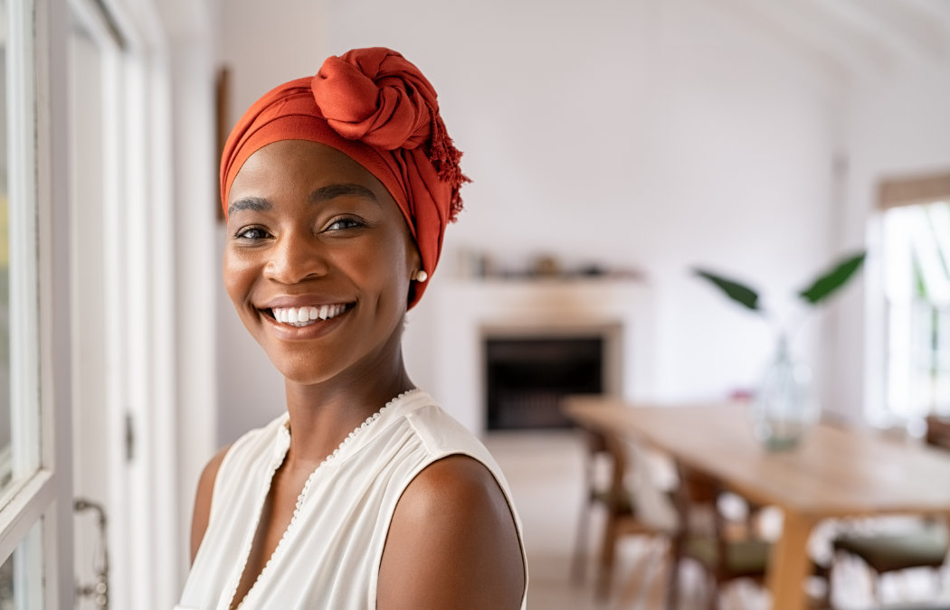 Smiling person wearing a red headwrap stands in a bright room with a wooden dining table and plants in the background.