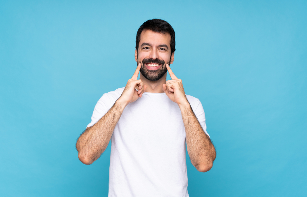 A man in a white t-shirt smiles against a blue background, pointing at his smile with both index fingers.