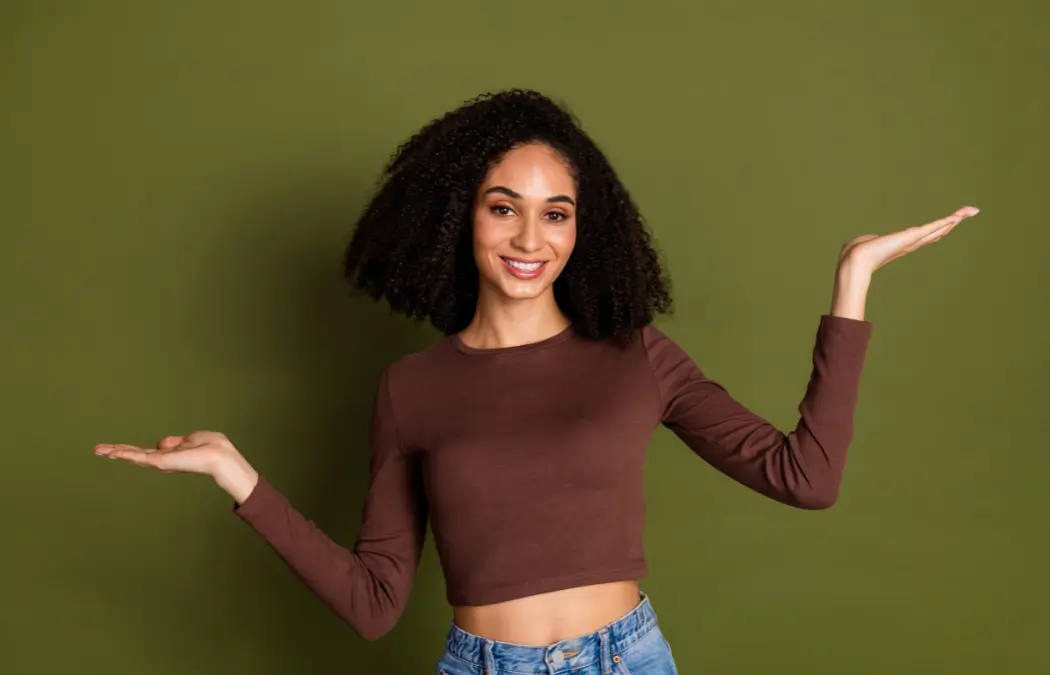 A person with curly hair in a brown shirt and jeans poses against a green background, smiling and holding both arms up as if balancing something.