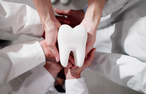 A group of people in white coats holds a large white tooth model together, symbolizing teamwork or collaboration in dentistry.