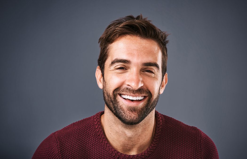 A man with short brown hair and a beard smiles against a gray background while wearing a maroon sweater.