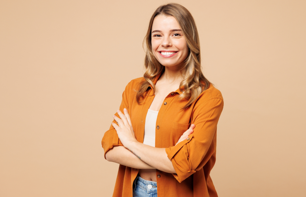 Person with long hair smiling, wearing an orange shirt, arms crossed, standing against a beige background.