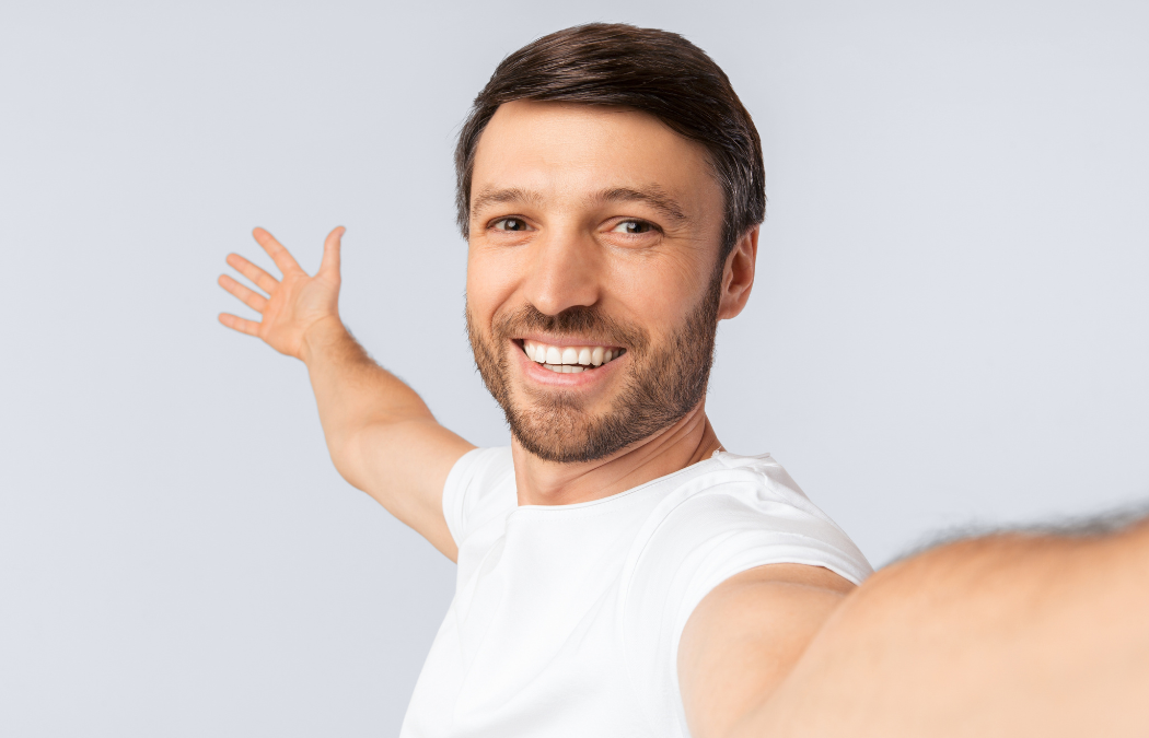 A man in a white shirt takes a selfie with one arm outstretched, smiling at the camera against a plain background.