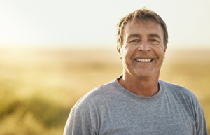 A man in a gray t-shirt smiles at the camera, standing outdoors with a blurred natural background.