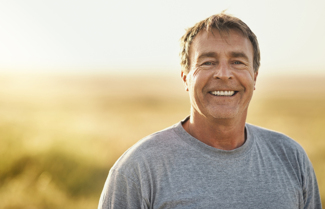 A man in a gray t-shirt smiles at the camera, standing outdoors with a blurred natural background.