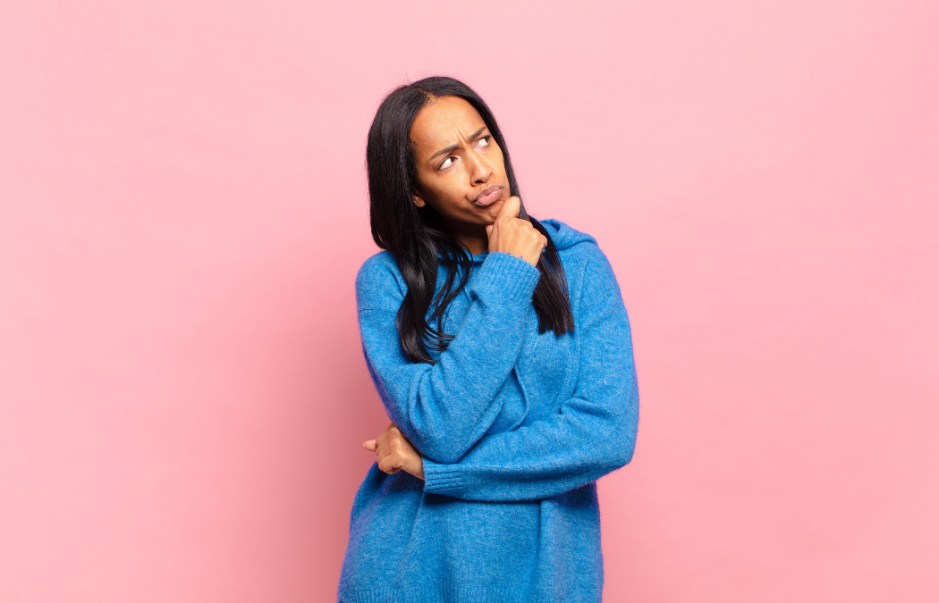 Person in a blue sweater with long dark hair, posing thoughtfully against a pink background.