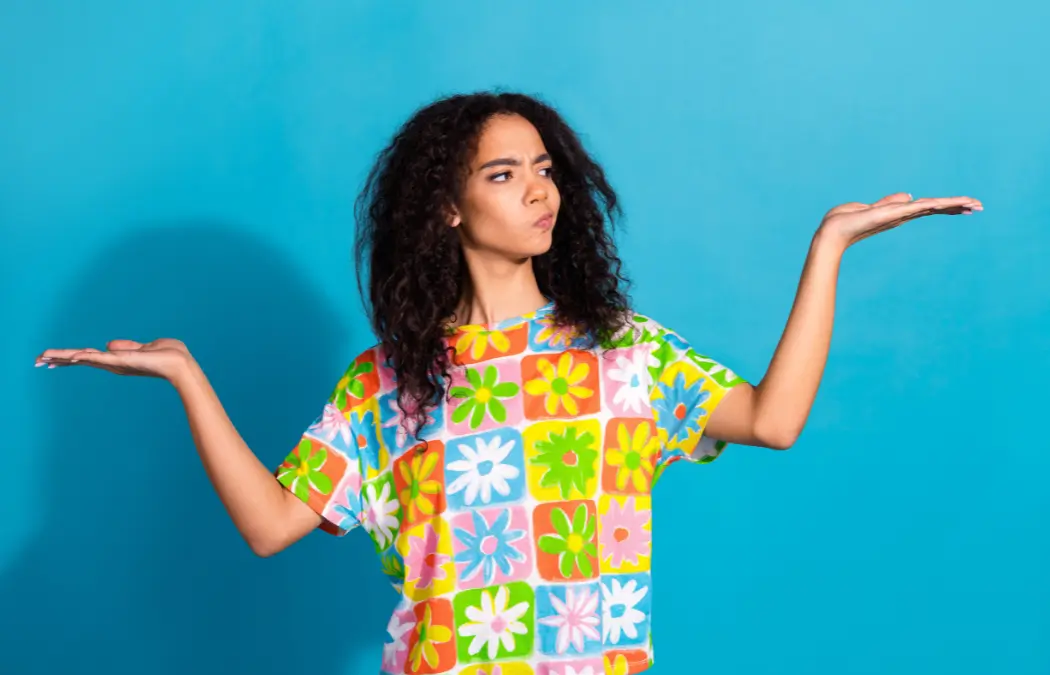 Person with curly hair wearing a colorful floral shirt looks perplexed, holding both hands up against a blue background.