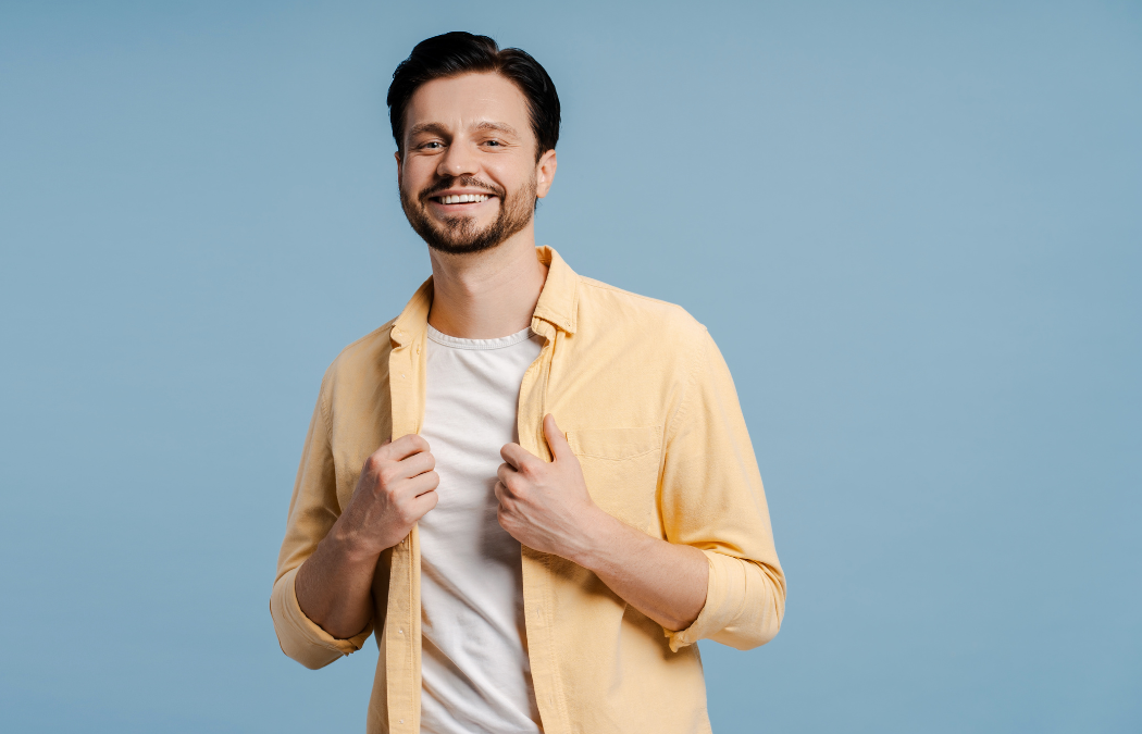 Smiling man in a yellow shirt and white t-shirt stands against a blue background, holding his shirt collar.