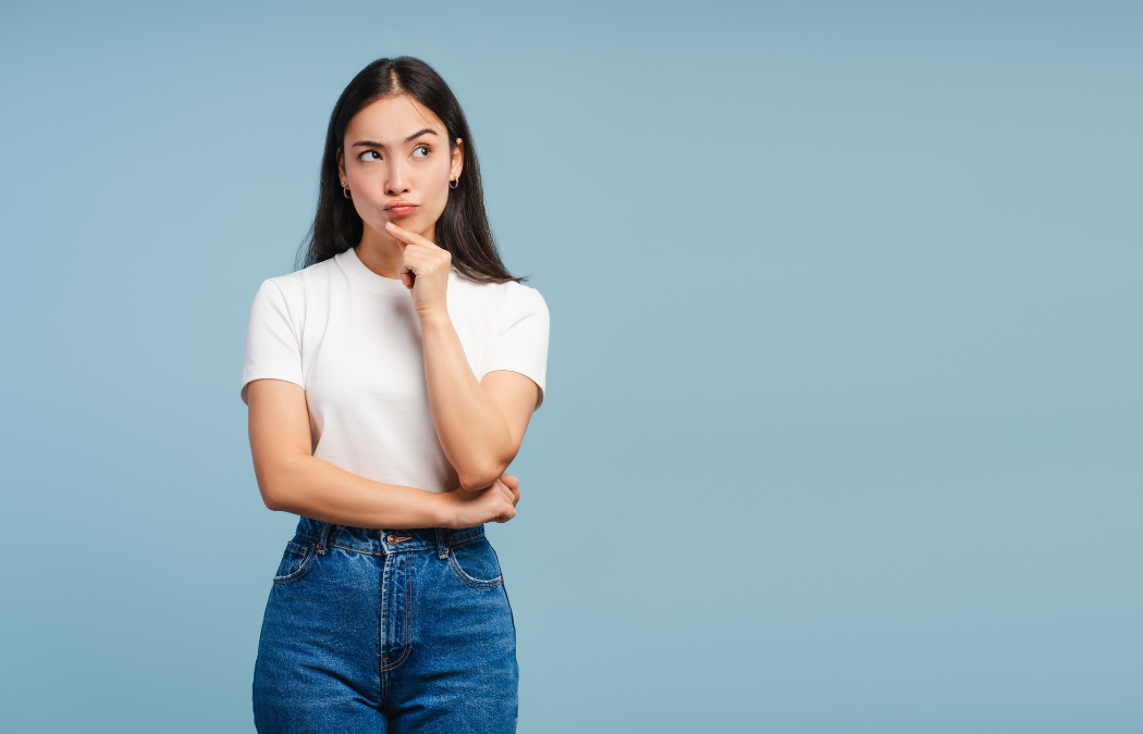 A woman in a white shirt and jeans stands against a blue background, looking up thoughtfully with her hand on her chin.