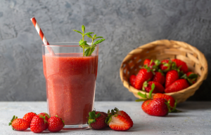 A glass of strawberry smoothie with a striped straw and mint garnish is on a table. Fresh strawberries are scattered next to a woven basket in the background.
