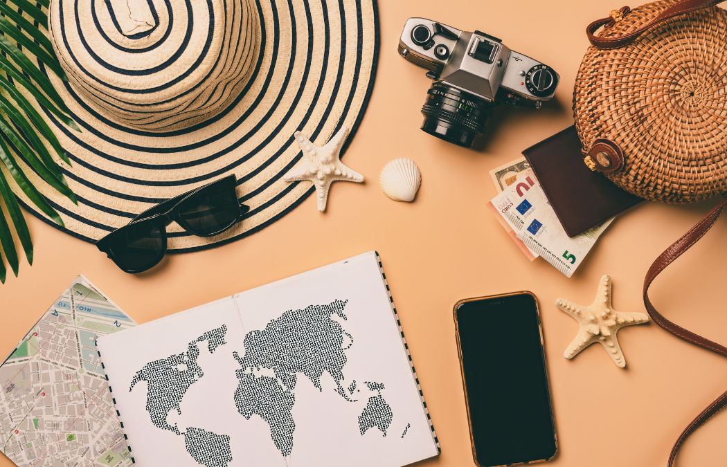 Travel essentials with a hat, camera, rattan bag, sunglasses, map, passport, currency, starfish, and a smartphone on a peach background.