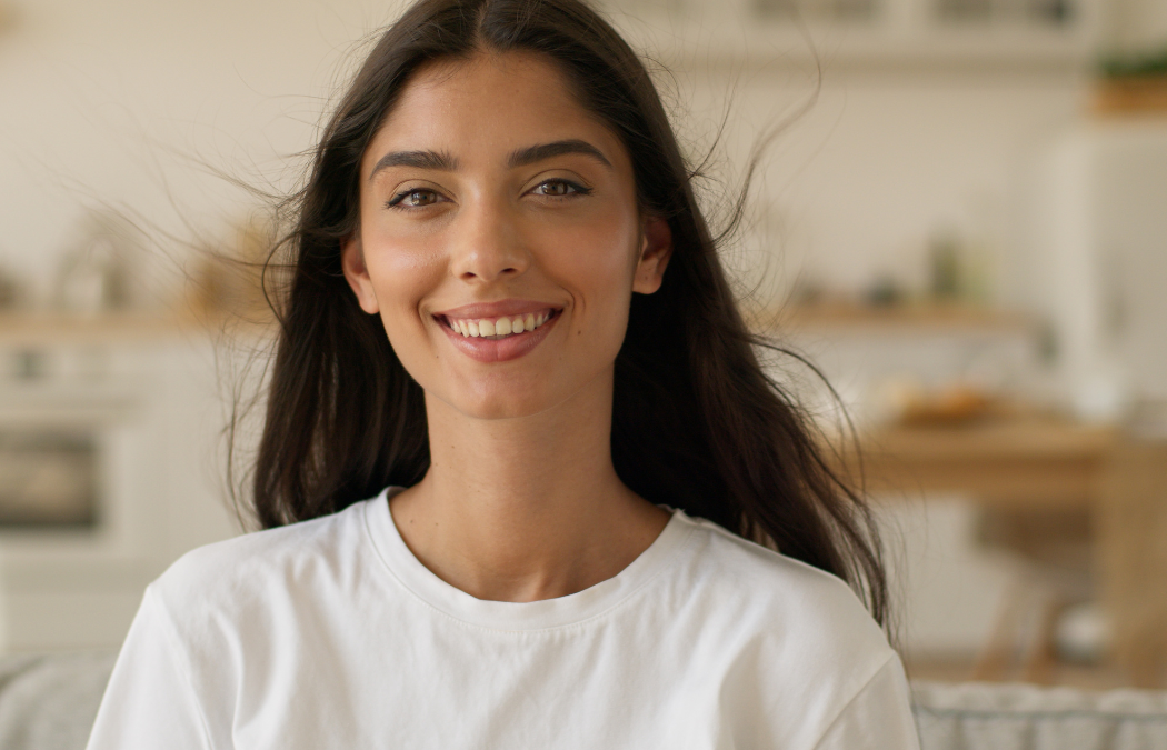 Person with long dark hair smiling, wearing a white shirt, in a bright indoor setting.